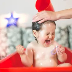 Małe dziecko panicznie boi się mycia głowy, krzyczy i płacze, gdy mama polewa mu włosy wodą z czerwonego kubeczka.