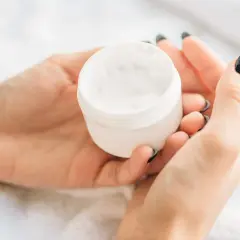 Dłonie trzymają biały słoiczek z kremem, w którym znajduje się dehydroacetic acid w kosmetykach.