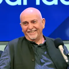 Uśmiechnięty Peter Gabriel, legendarny wokalista, w studiu. W tle niebieskie logo, przypominające o jego bogatej karierze z zespołem Genesis.