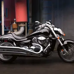 Błyszczący, czarny Suzuki Intruder 1800 z chromowanymi elementami, zaparkowany na tle industrialnej ściany z rurami.