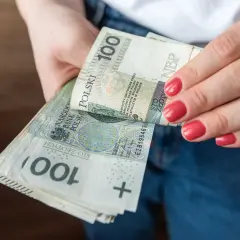 Dłonie trzymające stos banknotów 100 zł. Porównanie zasiłku stałego a renty socjalnej.