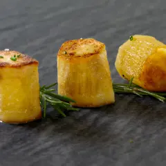 Drei goldbraune Fondantkartoffeln, garniert mit Rosmarin und Schnittlauch, auf dunklem Stein serviert.