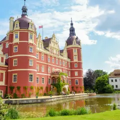 Czerwony zamek z wieżami, otoczony zielenią i wodą, stanowi serce mużakowskiego parku krajobrazowego.