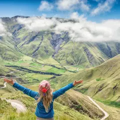 Mujer con brazos abiertos admira un paisaje montañoso verde, como si estuviera en la cima de la piedra del molino, con nubes y un camino sinuoso.