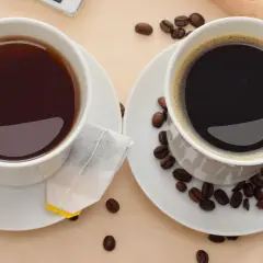 Zwei Tassen, eine mit schwarzem Tee und Teebeutel, die andere mit Kaffee und Kaffeebohnen. Perfekt, um Kaffee und Tee zusammen zu trinken.