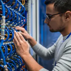 Specjalista w okularach pracuje przy serwerach, porządkując kable. To praca w dziedzinie teleinformatyki, kluczowa dla działania sieci.