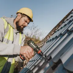 A roofer in a yellow hard hat and hi-vis vest uses a drill for metal roof repair.
