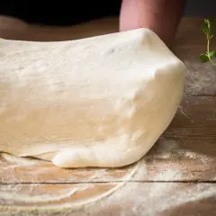 Frischer Teig für Pizza Semola wird auf einem Holztisch geknetet, bestäubt mit Mehl. Kräuter liegen daneben.