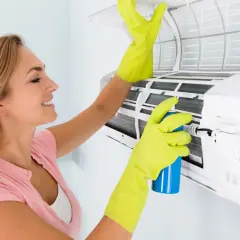 Mujer con guantes verdes limpia el aire acondicionado que huele a vinagre, rociando un producto para eliminar olores.