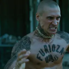 Wściekły mężczyzna z tatuażem "Furioza" na klatce piersiowej, w złotym łańcuchu, w tle druga postać z bronią.