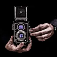 Dwie dłonie trzymają zabytkowy aparat Rolleiflex, którego obiektywy odbijają światło, tworząc wrażenie złotych proporcji w kadrze.
