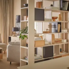 Przytulny salon z biurkiem, podzielony regałem z książkami i dekoracjami. Jasne meble i zasłony tworzą ciepłą atmosferę.
