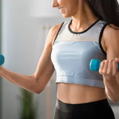 Uśmiechnięta kobieta wykonuje ćwiczenia na biceps w domu, podnosząc niebieskie hantle.