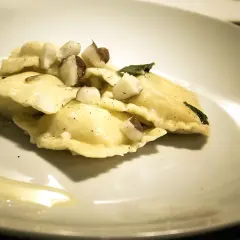 Ravioli pere e pecorino, conditi con nocciole tritate e una foglia di salvia, su un piatto bianco con un filo d'olio.