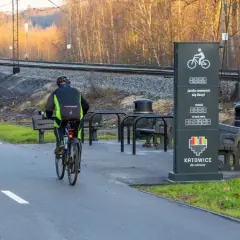 Rowerzyści korzystają z nowoczesnych tras rowerowych w Gliwicach. Obok torów kolejowych, przy stojaku z licznikiem przejechanych kilometrów, panie i panowie cieszą się aktywnością na świeżym powietrzu.