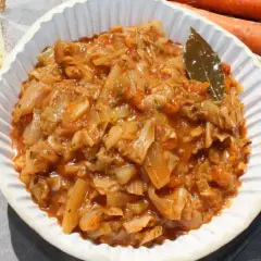 Pyszny bigos z młodej kapusty w białej misce, obok kromka chleba i marchewki.