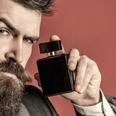 Mężczyzna z brodą trzyma czarną buteleczkę perfum na czerwonym tle.