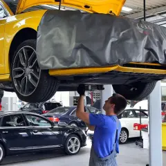 Mechanik pracuje pod żółtym Audi, sprawdzając, ile kosztuje wymiana oleju silnikowego w samochodzie. W tle inne auta.