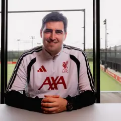 Uśmiechnięty trener Liverpoolu w stroju treningowym z herbem klubu i logo AXA. Za nim boisko treningowe.