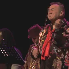 Michał Olczak gra na skrzypcach, ubrany w kolorową koszulę. Obok niego muzycy z dredami i w kapeluszu.