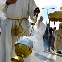 Procesja Bożego Ciała z kadzidłem i monstrancją. Uczestnicy w białych szatach idą ulicą.
