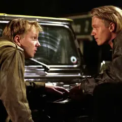 Dwóch młodych mężczyzn w skórzanych kurtkach, jak z filmów o mafii, pochyla się nad maską samochodu, prowadząc intensywną rozmowę.
