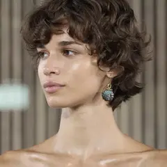 Mujer con pelo corto fino y escaso, rizado y castaño. Luce un pendiente colgante con una piedra grisácea.