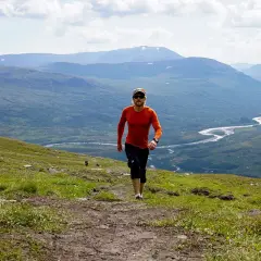 Man springer utför en grön sluttning med en slingrande älv i bakgrunden, en vy över Blåhammaren och Storulvån.