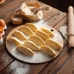 Surowe pierogi ułożone na desce, obok jajka, mąka i wałek.