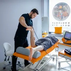 Fizjoterapeuta wykonuje zabieg falą uderzeniową na stopę pacjenta.