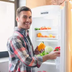 Hombre sonriente saca pimientos de la nevera. Se pregunta cuánto dura el pisto en la nevera, mientras organiza sus verduras.