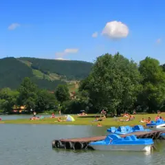 Słoneczny dzień nad jeziorem w Międzybrodziu Żywieckim. Ludzie odpoczywają na plaży, a przy brzegu cumują niebieskie rowerki wodne.