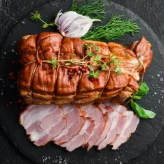 Wędzenie szynki na gorąco: apetyczny, brązowy kawałek mięsa, ozdobiony ziołami i pieprzem, obok plastrów soczystej szynki.