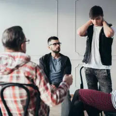 Grupa ludzi siedzi w kręgu, jeden mężczyzna stoi, wszyscy wydają się słuchać.