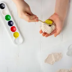 Kind bemalt Gipsfisch mit gelber Farbe. Wasserschale und Farbpalette daneben.