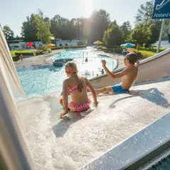Kinder rutschen im Freibad Leutkirch auf einer Wasserrutsche. Im Hintergrund das Schwimmbecken mit Badegästen.
