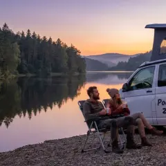 Para relaksuje się przy ognisku podczas **camping al lago**. W tle spokojne jezioro i las o zachodzie słońca.