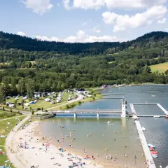 Słoneczny dzień nad zalewem Stara Morawa. Ludzie odpoczywają na plaży i w wodzie, a w tle widać las i pola namiotowe.