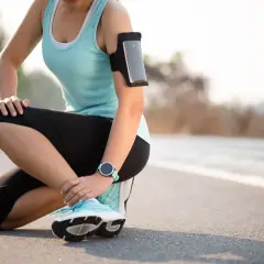 Kobieta w stroju sportowym klęczy na drodze, trzymając się za bolącą kostkę.