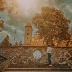 Osoba na schodach przed kamiennym murem z godłem Polski, w tle kościół w Czerwińsku nad Wisłą.
