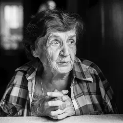 Portret starszej kobiety w kratę, z dłońmi złożonymi na stole. Obraz jest czarno biały, pełen zmarszczek i historii.