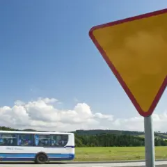 Żółty trójkątny znak a7 ostrzega przed ustąpieniem pierwszeństwa. W tle jedzie autobus, a za nim zielone wzgórza.