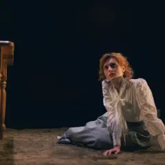 La signorina Julie, con capelli rossi e un vestito bianco, è inginocchiata su un pavimento di terra, con uno sguardo perso nel vuoto.