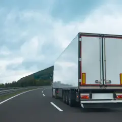 Ciężarówka na autostradzie, z tyłu widać białą naczepę. Na poboczu rosną drzewa, a nad nimi chmury. Tablice wyróżniające przepisy są widoczne na naczepie.