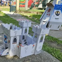 Zabawki z kartonu diy: biały zamek z wieżami i schodami, obok rakieta. Dzieci bawią się w tle.