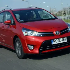 Czerwona Toyota Corolla Verso jedzie po drodze. Kierowca w okularach przeciwsłonecznych prowadzi auto.