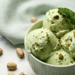 Coppa di cremoso gelato al pistacchio, guarnita con foglioline di menta e granella di pistacchi. Alcuni pistacchi interi sparsi sul marmo.