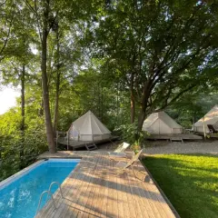 Luksusowy glamping nad jeziorem. Białe namioty na drewnianych tarasach, basen i leżaki wśród zieleni.
