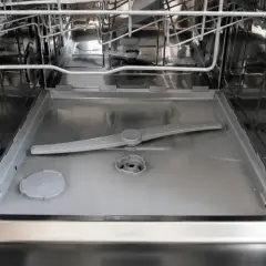 Intérieur d'un lave-vaisselle ouvert, montrant le bras d'aspersion et le fond. On observe de l'eau stagnante au niveau du filtre, signe d'un nettoyage nécessaire.
