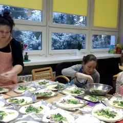 Trzy kobiety w fartuchach przygotowują jedzenie na stole, na tle okien ze śniegiem.